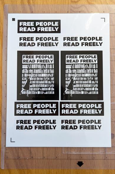 A sheet of stickers reading “Free People Read Freely” in both black on white and white on black, and a larger sticker with the same text, above a print of a person in a dress reaching to take a book from a large bookshelf