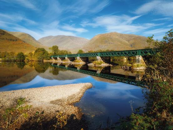 A Victorian-era railway bridge, painted in deep forest green, spans gracefully across Loch Awe, its latticed iron framework and stone pillars casting perfect reflections in the still waters beneath. The bridge is beautifully framed by the autumnal Scottish Highlands, their ochre slopes rising majestically against a brilliant blue sky streaked with wispy clouds, whilst a small gravelly peninsula juts into the loch's crystalline waters in the foreground.