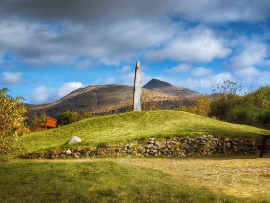 A tall granite memorial stands atop a grassy knoll, backed by the imposing peak of Ben Cruachan beneath a dramatic sky of racing clouds, whilst a rustic stone wall wraps around the mound's base and a wooden bench sits quietly to the left, all framed by autumn-touched foliage and the golden Highland light illuminating the verdant grass.