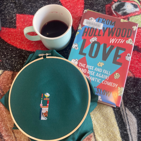 A mug of tea, a copy of the book "From Hollywood With Love: The Rise and Fall (And Rise Again) of the Romantic Comedy, and a cross stitch in a wood embroidery hoop.