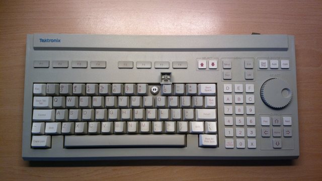 Eine graue Tektronix-Tastatur mit hellgrauen Modifier-Keys und braunroter Beschriftung. In der untersten Tastenreihe hat es nur Caps-Lock, eine 10.5U Leertaste und PrintScreen. Die Funktionstasten (nur F1 bis F8) sind kleine, flache, rechteckige Buttons, die ca. doppelt so breit wie hoch sind.

Die Tastenkappe der Taste "9" ist abgenommen und den Switch-Mount darunter zu zeigen.

Anstatt dem MF-II- und dem typischen Nummernblock hat sie unter anderem (innen) einen Ziffernblock mit folgender Tastenanordnung:

D E F
A B C
7 8 9
4 5 6
1 2 3
H 0 X (hier steht das "H" für "HELP NOTES")

Rechts außen ist ein Block mit kreuzförmig angeordneten Cursor-Tasten und einem drei Tasten breitem Drehrad mit Fingerloch und Rändelung.

Darüber noch drei Tasten "Start/Stop", "Auto" und "Cont".