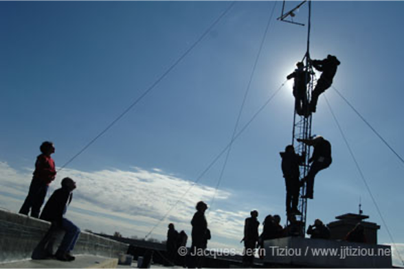 People on a roof against a blue sky, watching and holding cables, while a few other people climb an antenna tower mounted on the roof