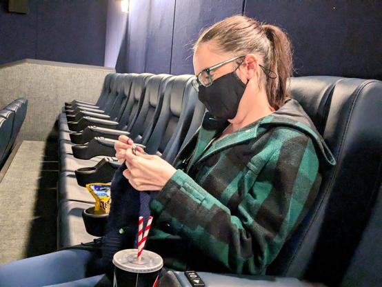 Photo of a white woman with brown hair tied back, sitting in a cinema seat in an otherwise empty row. She's wearing a green and black checkered coat, and a black facemask. She's also focused on some knitting although the light in there is so bad it's difficult to make out the actual project (it's a navy jumper).