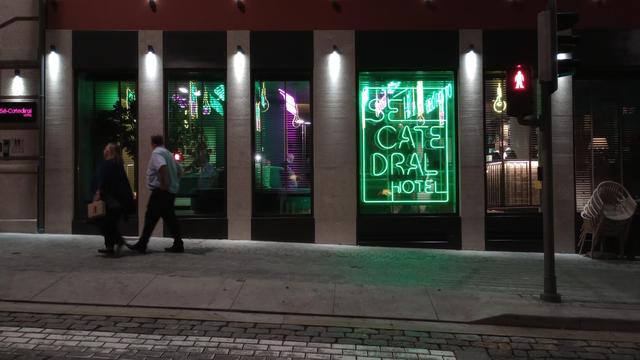 Nacht. Erdgeschoss-Fassade eines Hotels mit einer neongrünen Leuchtreklame die den Namen des Hotels "Sé Catedral Hotel" zeigt. Die Gebäudefront wird durch graue, mit Strahlern beleuchtete Säulen zwischen den Fenstern unterteilt. Zwei Menschen schlendern vor dem Hotel über den grau gepflasterten Gehsteig.