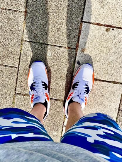 Camera pointing down on sneakers while walking on a pedestrian path, showing Nike Air Max 90 in white with orange and blue accent colours.