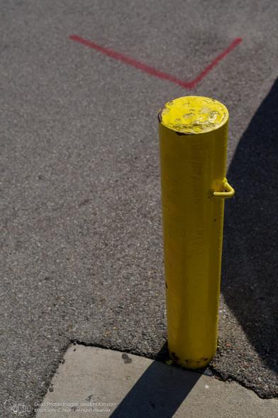 A photograph of a cylindrical iron stanchion filled with concrete and painted yellow. It has a triangular hook eyelet welded to the side. Beyond, a red line painted at a fight angle on frey asphalt appears to point at the top of the stanchion, In the foreground, a shadow falls across a triangle of light-grey concrete.