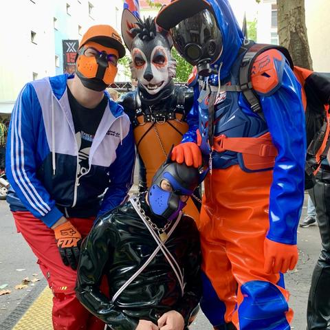 Four gearheads: To the left the fox in his orange puppy hood, a blue-white Adidas windbreaker jacket and red e.s. motion pants; in the middle PupZuki in his Spikey boy rubber pup hood and black-orange rubbersuit; to the right Adensy in shiny orange-blue raingear, matching his Fox MX protector and gas mask; and in the middle front, kneeling, xolo_93 in his blue puppy hood and black rubbersuit, wearing a lock chain.