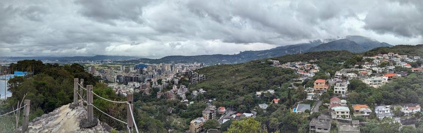 A view over northern parts of Taipei, Taiwan from near a hill called Battleship Rock. You see small houses, highrises and a sports field against a backdrop of hills and low clouds covering the top of these hills.