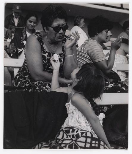 The image is a black-and-white photograph depicting several individuals seated closely together, likely at an event such as a sports game or concert. In the foreground, there's a woman wearing sunglasses and dark clothing with floral patterns; she appears to be in her middle years, engaged in conversation while holding what seems like food wrapped in napkins between fingers of one hand.

Behind her is another person seemingly smoking a cigarette. The central figure has their hair styled in braids or twists that reach down the back of their head and are held together with pink-colored bands; they seem to be younger than those around them, possibly a teenager or pre-teen girl who appears relaxed as she looks away from the camera.

Other individuals can also be seen seated behind this main group. They exhibit casual clothing styles typical for an informal outdoor event. Some have on patterned dresses and others in striped shirts; one is wearing sunglasses similar to the woman with food, indicating it might indeed be a sunny day or bright lighting condition within which the photo was taken.

The overall atmosphere seems lively but subdued as none of these individuals are displaying extreme emotions such as laughter or surprise visible.