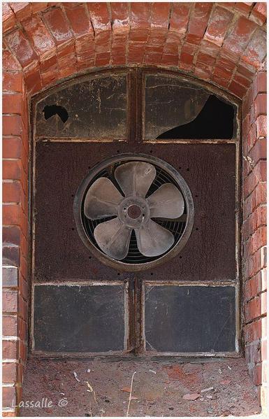Zu sehen ist ein Fenster im Erdgeschoss eines Ziegel-Fabrikbaus. Die Fensterfläche besteht aus einer verrosteten Stahlfläche, in die mittig ein Ventilator eingesetzt wurde, ober- und unterhalb jeweils zwei kleine, blinde Glasfenster. Die oberen sind teils zerbrochen.
Der Ventilator diente wahrscheinlich der Entlüftung des dahinter liegenden Raumes.