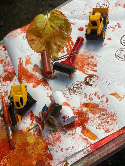 An art table in the aftermath. Red and yellow paints all over a roll of paper, with toy trucks, brayers, and leaves used to make marks.