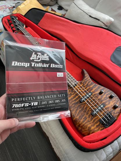 A photo of my hand holding a pack of LaBella “Deep Talkin’ Bass” 760FS-TB flatwound bass guitar strings. In the background, slightly out-of-focus, is my brown Peavey bass guitar lying in a plush gig bag with a red interior.