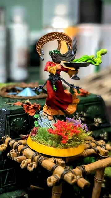Lumineth Realm-Lords Scinari Cathallar model from Games Workshop painted with a red dress with yellow trim.  The model is holding a silver chalice with neon green smoke billowing out; the model's face is covered in a dark grey mourning cloth.