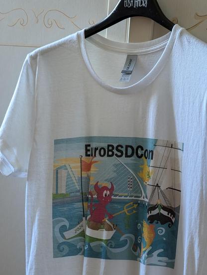 A white t-shirt with a colorful print featuring the EuroBSDCon 2024 logo, held in Dublin. The design shows a red FreeBSD daemon character (the 'Beastie') rowing a boat (with the NetBSD flag) in front of a cityscape with a bridge and a tall ship in the background. Puffy, the OpenBSD logo, is jumping. The print includes the EuroBSDCon 2024 text.