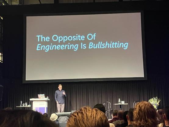 Alex Russel on stage. There is a big screen with text saying “The Opposite Of Engineering is Bullshitting”