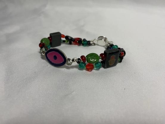 A colorful bracelet featuring various beads, including a large eye symbol, green and red elements, and a hand charm, all on a silver chain. The background is a smooth white surface.