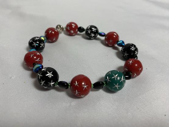 A colorful bracelet composed of round beads in red, black, and green, featuring metallic silver star designs. The beads are interspersed with small black and iridescent beads, displayed on a light background.