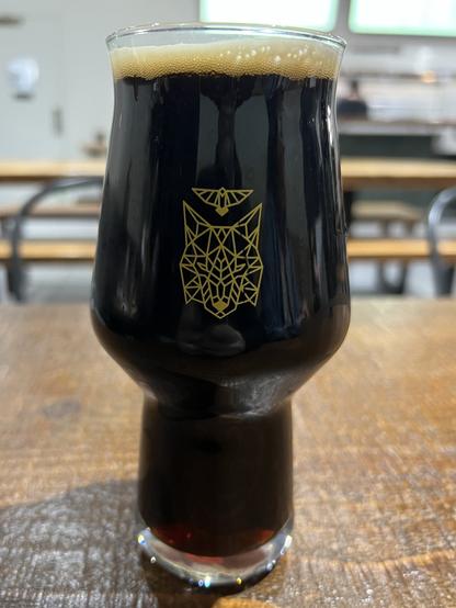 A glass with dark stout on a wood table top. Other tables and a restaurant pass through are unfocused in the background. The glass has a wireframe fox head with a stylized bird between its ears