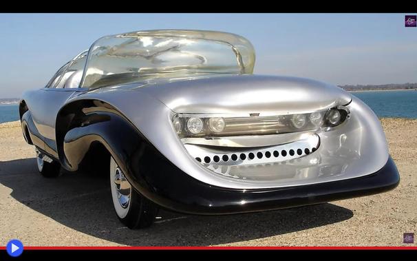 Automobile dalla strana carrozzeria color argento e nero, con abitacolo bulboso e trasparente che riflette lo scintillio di una spiaggia assolata. Il paraurti sporge vistosamente in avanti, come una sedia.