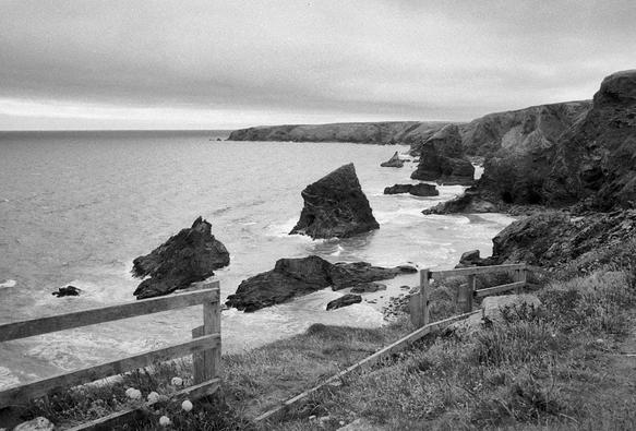 Black and white photo showing a Cornish coastal scene. There is a broken wooden fence in the foreground; through the gap can be seen several of the spectacular sea stacks called Bedruthan Steps, at high tide with the breaker around them. Near the top is a headland with moody sky above.