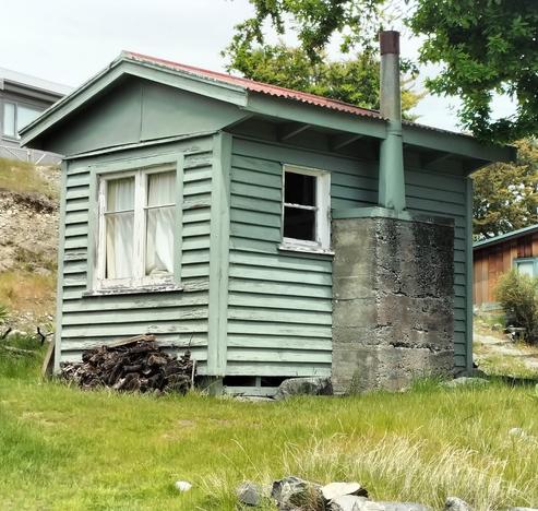 Very small one room weatherboarded Crib( or Batch) . One wall has a concrete fireplace and chimney. Grass and rocks about it.