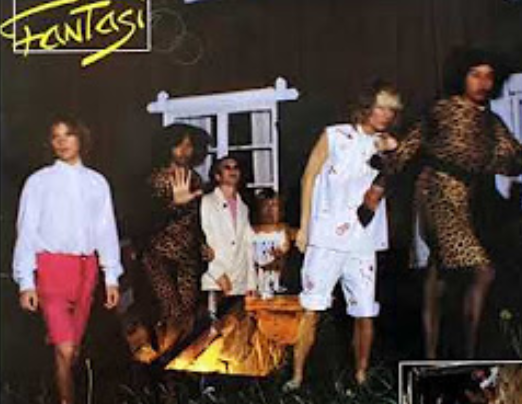 A low-res image of 6 people standing in front of a window around a fire, dressed in eighties outfits. The text 'fantasi' is written in the top-left corner in a handwriting-style yellow font.
