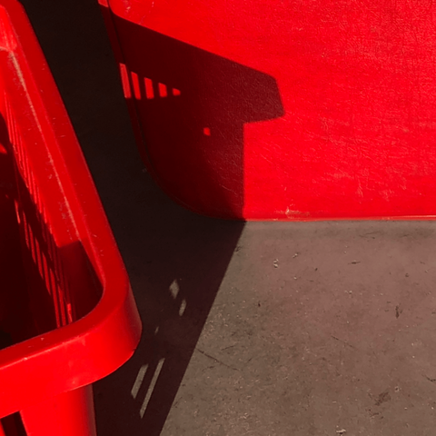 Corner of a red plastic shopping basket seen on the left of the frame, sitting on a bare concrete floor, casting a shadow against the side of a bright red suitcase.