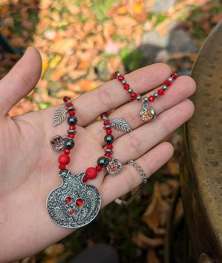 A handmade necklace with 3 pomegranate pendants (one large two small), red and hematite styled beads, sterling silver leaf shaped pendants and a fitting beaded bracelet with a pomegranate pendant 
On the palm of my hand with fall leaves in the background