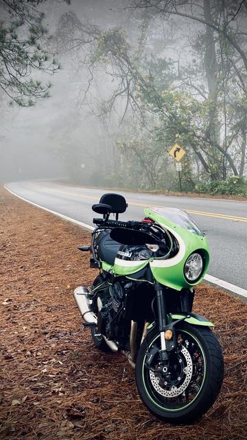 Bayport's green and black Kawasaki z900rs-cafe parked on the shoulder of a forest road. In the background is a road sign indicating a curve ahead and the forest is shrouded in foggy fog.