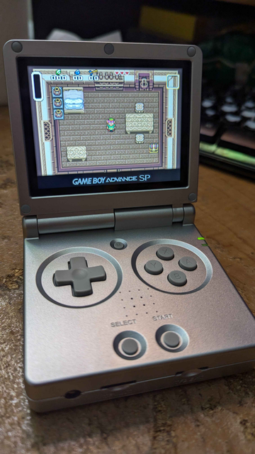 A retro emulation handheld device that looks remarkably similar to a silver Gameboy Advance SP. It is a clamshell device that closes. The game being displayed on the screen is The Legend of Zelda: A Link to the Past, the GBA version.