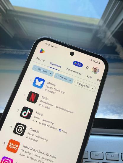 Photo of the top-free-apps chart in the U.S. Play Store, as seen in a Pixel 8 phone held above a laptop screen showing a picture I took of intersecting contrails as seen from a flight between Houston and Austin. You can see a window above the phone, with a blue sky visible through the skylight.