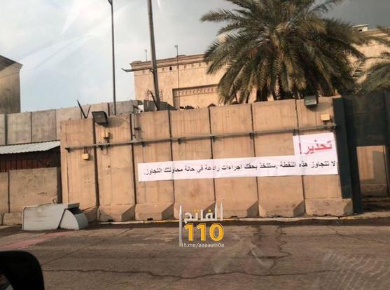 A warning sign is displayed prominently on a concrete wall, stating "لا تتجاوز هذه النقطة، ستتخذ بحقك إجراءات رادعة في حالة تجاوزك" (Do not cross this point; strict measures will be taken against you if you do). The background features palm trees, adding to the outdoor setting.