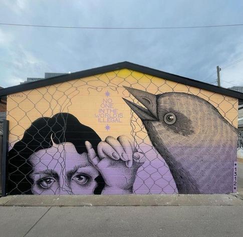 Streetartwall. An impressive mural with a message has been painted on the outside wall of a flat house. The wall is divided into the colors light yellow (top) and purple (bottom) and shows the head of a dark-haired girl behind a wire fence. The fingers of her hand are clutching the wire. The head of an oversized gray bird is also pecking at the wire from the right, creating a hole in the fence. In the center above this hole is written in purple: "No one in the world is illegal" The mural was created by several artists in unusual painting styles. The girl is reminiscent of Art Nouveau ladies and the large bird is reminiscent of charcoal drawings from old zoology books. Beautiful.
Info: The Artists: "A reminder to blame governments, landlords and billionaires for todays crisis, not immigrants".