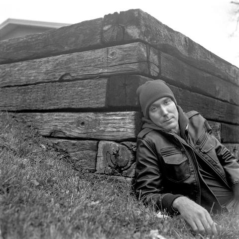 Black & White photo of a man in a leather jacket is laying down and leaning against a wooden retaining wall on slope.