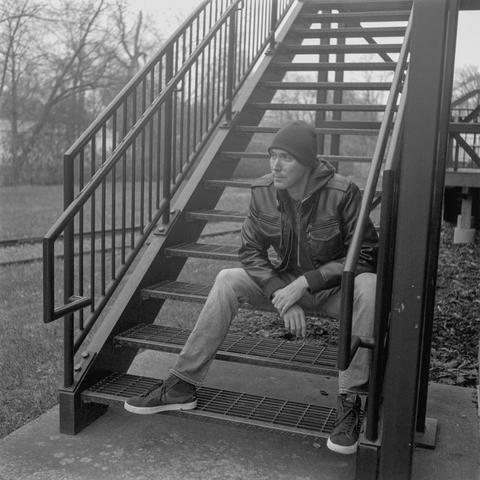 Black & White photo of a man in a leather jacket sitting on some metal stairs looking off camera.