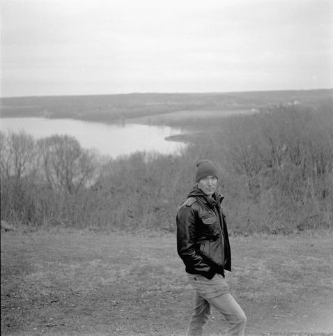 Black & White photo of a man in a leather jacket posing against a backdrop of a river valley looking into the camera.