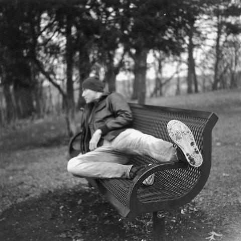 Black & White photo of a man in a leather jacket laying on a park bench with outstretched shoes which is what is in focus.