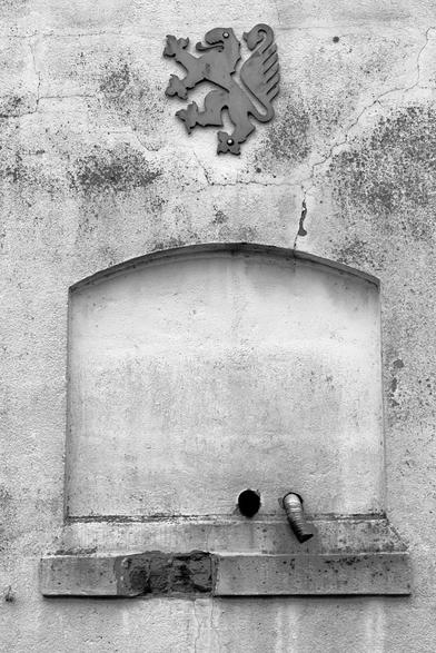 A bricked-up window. Two holes have been made in the wall, from which two drainpipes lead out. A piece of brickwork has broken out of the window sill on the left. Above everything, someone has attached the lion coat of arms of Brunswick. The picture is in black and white.

##

Ein zugemauertes Fenster. Durch die Mauer wurden zwei Durchbrüche erstellen aus denen zwei Abflussrohre nach außen führen. Aus dem Fenstersims ist auf der linken Seite ein Stück Mauerwerk herausgebrochen. Über allem hat jemand das Löwenwappen von Braunschweig angebracht. Das Bild ist in schwarzweiß.