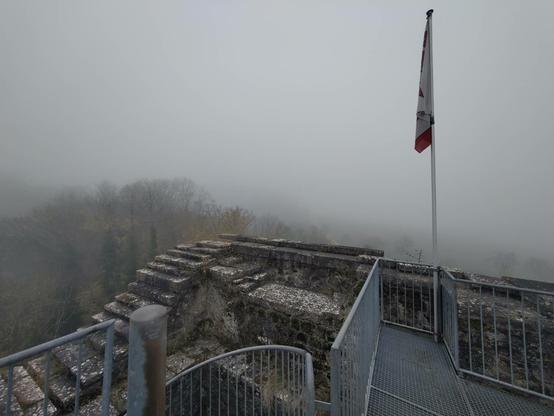 Aussicht von einem alten, gemauerten Turm mit Metallgeländer und einer schlaffen Fahne an einem Mast. In der Umgebung ist etwas Wald zu sehen, der Rest ist nur nebelgrau.
