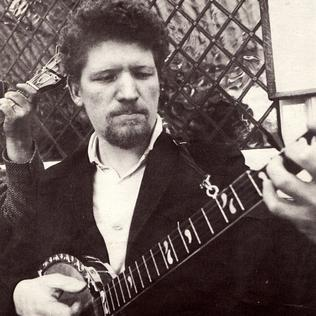 Black and white picture of Luke Kelly (with focus on torso, not a full length picture). He has goatee (beard) and curly hair, and is playing a banjo.