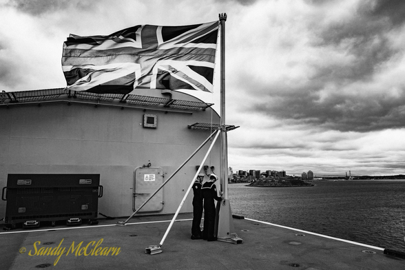 Sailors raising the Union Jack on an aircraft carrier with a cityscape in the background.