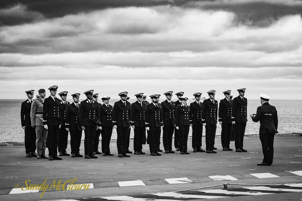 Officers at attention on deck with clouds behind.