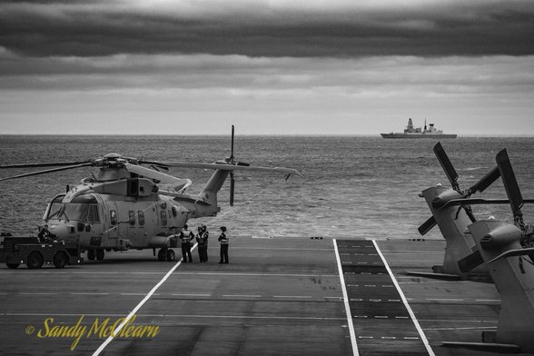 Helicopters stowed on deck with a destroyer on the horizon.