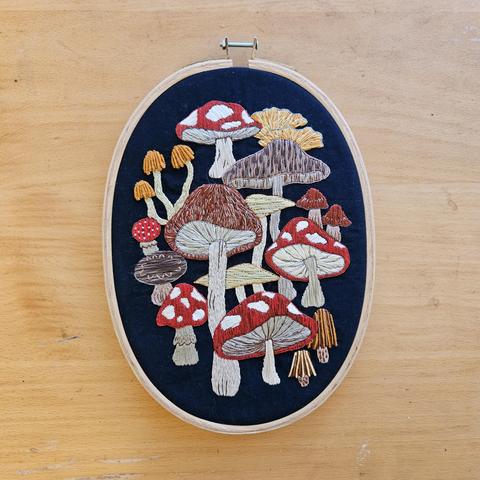 An oval embroidery hoop containing a mushroom themed design, depicting a diverse array of fungi on a navy blue background.
