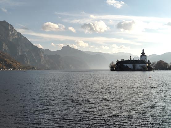 Blick hinaus auf den See. Dahinter einige felsige Berge, im See auf einer kleinen Insel erbaut ist das Schloss Orth mit einem kleinen Turm, von rechts führt eine Brücke über den See zum Schloss. Blauer Himmel mit ein paar vereinzelten Wölkchen.