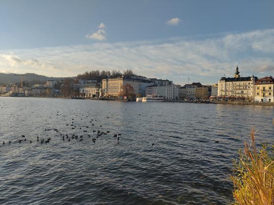 Blick über den See hinüber auf Gmunden. An der Seepromenade stehen einige schöne historische Gebäude, dahinter zieht sich die Stadt Gmunden über den Hang hinauf.