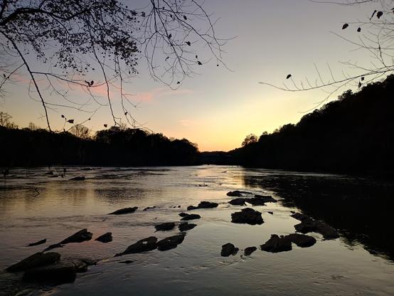 Post sunset glow over a river. Mostly dark blues and yellows. A little bit of red/pink reflected in some small whispy clouds. The trees and bluff on the far bank are black against the evening light.