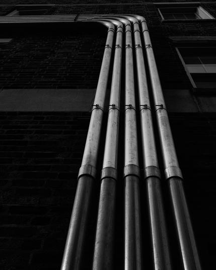 The image shows power conduits affixed to a brick wall in a back alley. The conduits have a metallic sheen and cast distinct shadows on the brick surface, creating a play of light and dark, highlighting their shapes against the masonry.