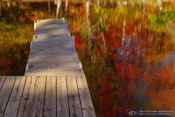 A photograph of an L-shaped dock with weathered wooden planks and white mooring cleats. The dock extends from the lower left of the frame into water that reflects the trunks and colorful leaves of an autumn forest. The water has slight ripples on its surface, which makes the forest look a little distorted or impressionistic.