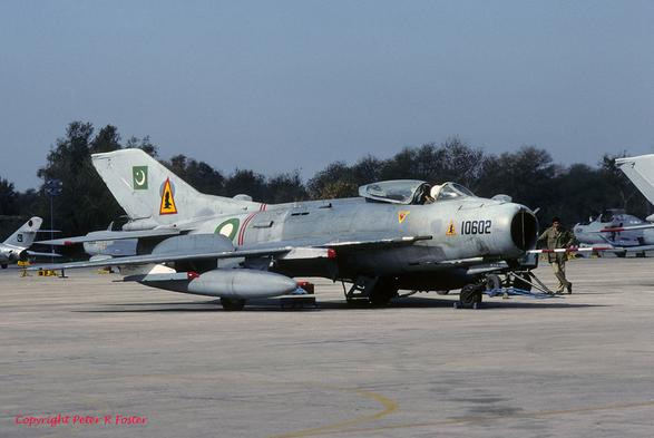An F-6 of Pakistani Squadron 25 parked on the apron of Rafiqui Airbase, Punjab, Pakistan on 14 Feb 1991. A pilot wearing a white helmet monitors cockpit interments during startup. The canopy of the cockpit is slid back open. A member of the ground crew in a green uniform stands lazily beside an external power unit parked beside the nose of the aircraft.